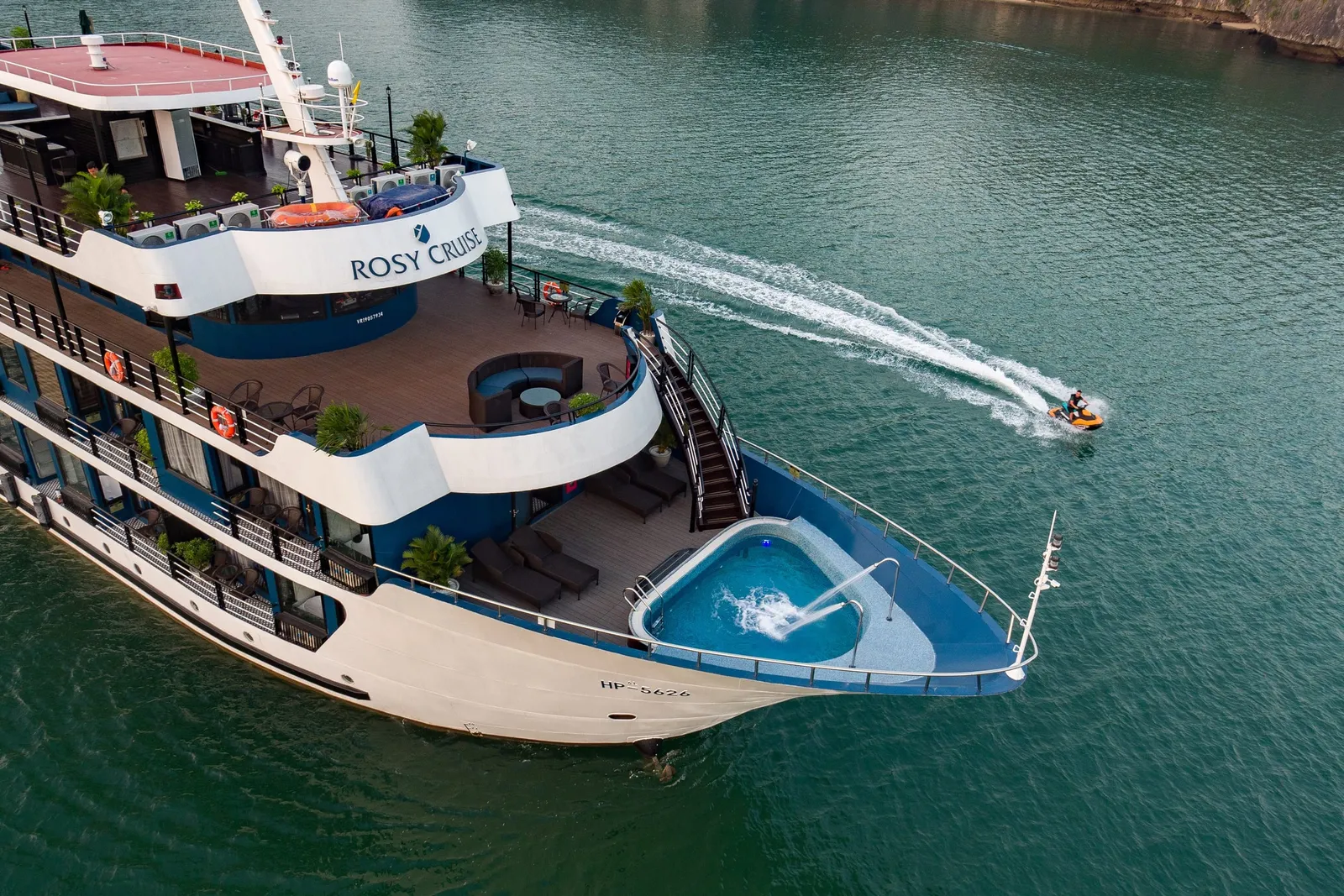 Aerial view of a multi-deck white and blue cruise ship with 'Rosy Cruise' on its side and a bow plunge pool, while a person on a jet ski leaves a wake in green water near a rocky cliff.