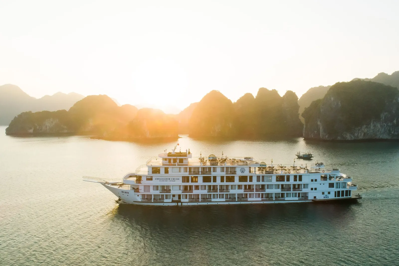 An aerial view of a large white multi-deck cruise ship with 'Ambassador Cruise' lettering on its hull, sailing on calm water past limestone karst islands in golden sunset light.