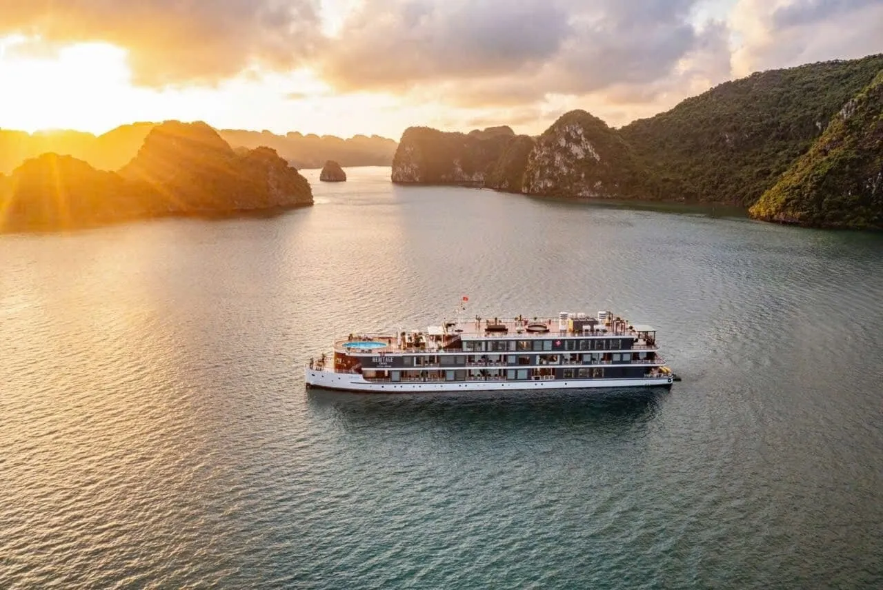 Aerial view at sunset of a multi-deck white cruise ship with a rooftop pool, with the name 'Heritage' on its side, sailing on calm water among green limestone karst islands.