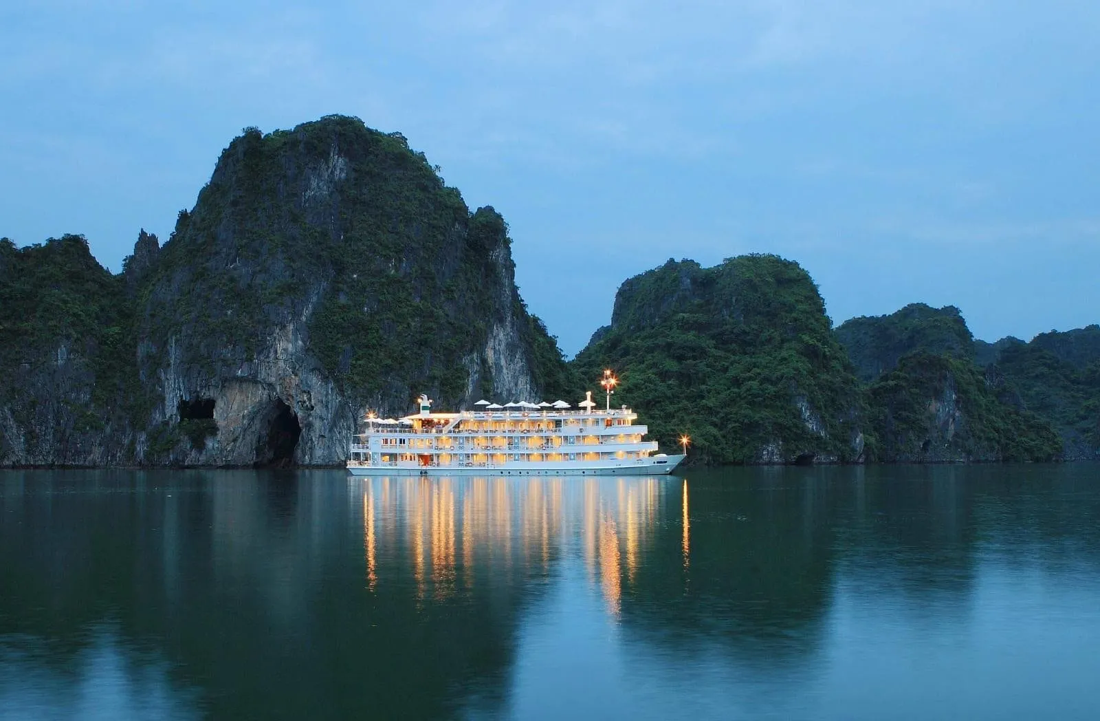 A multi-deck white cruise ship lit up at dusk sits on calm water in front of tall green limestone karst cliffs, with its lights reflected in the water.