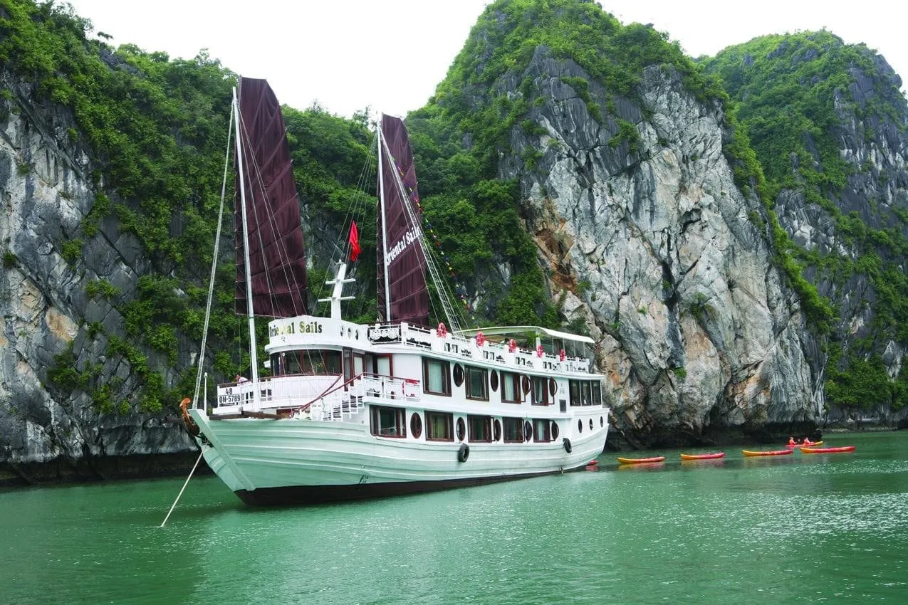 A white two-deck wooden junk with dark maroon sails is moored in green water beside limestone karst cliffs, with several red and yellow kayaks nearby.