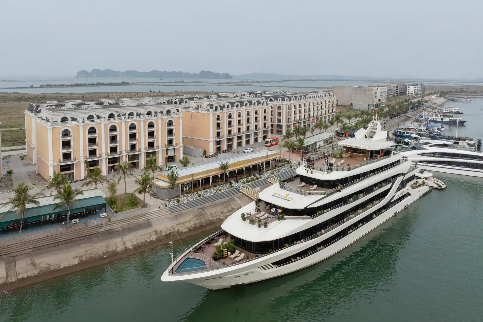 Aerial view of a large white multi-deck cruise ship with a yellow logo on its upper deck, moored at a quay beside a cream-colored neoclassical-style building complex, with limestone karst hills in the hazy distance.