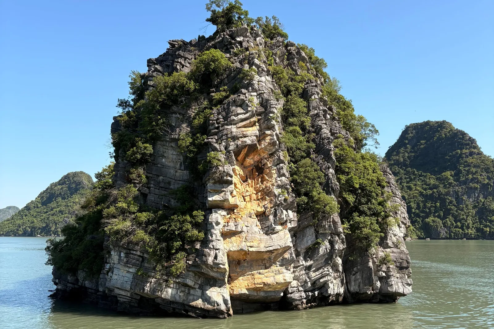 A vegetated limestone karst rock rising from green water under a clear blue sky, viewed from a boat deck with a red and metal railing and wicker furniture in the foreground.