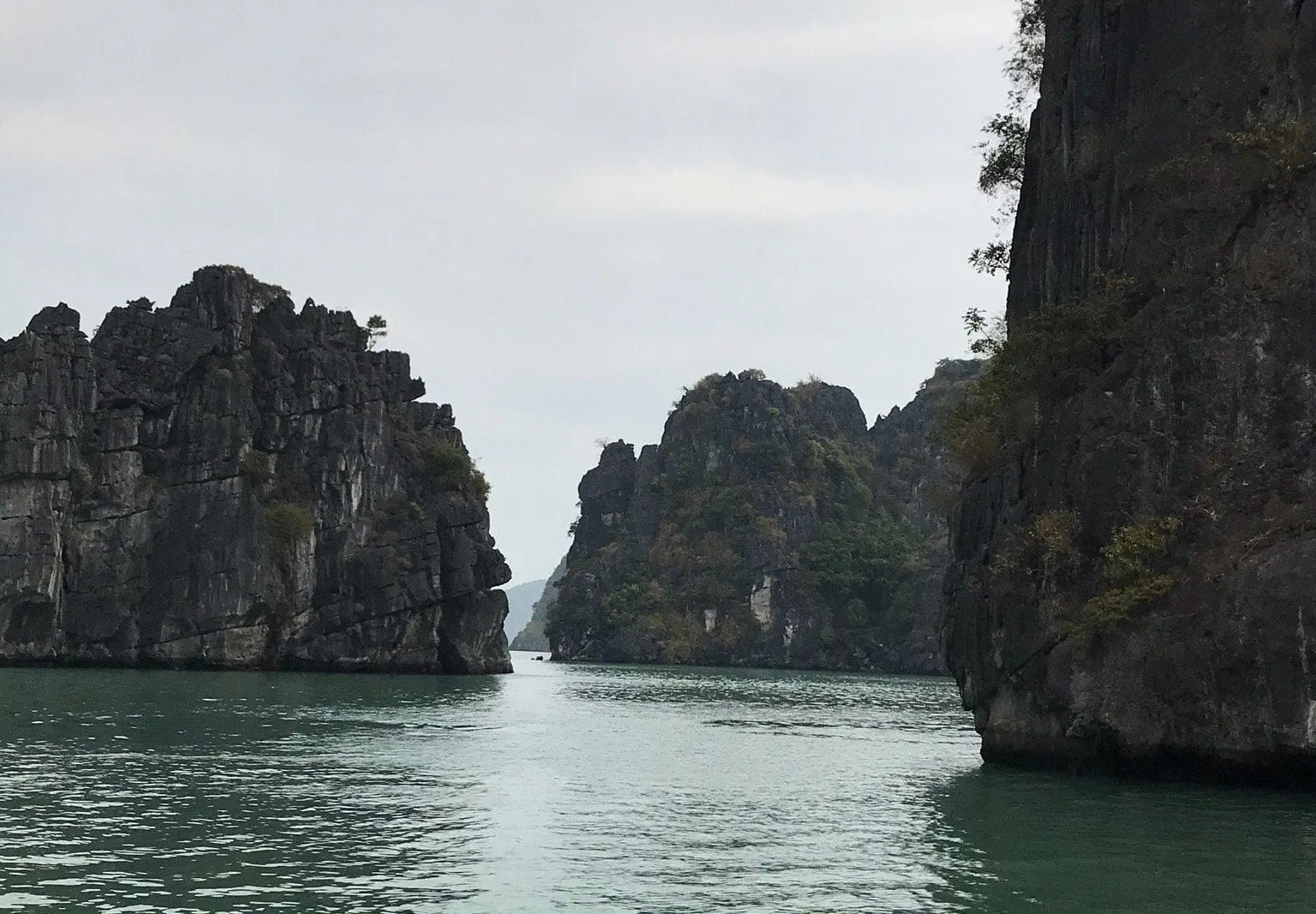 A narrow channel of green water between dark limestone karst cliffs under an overcast sky, photographed from water level.