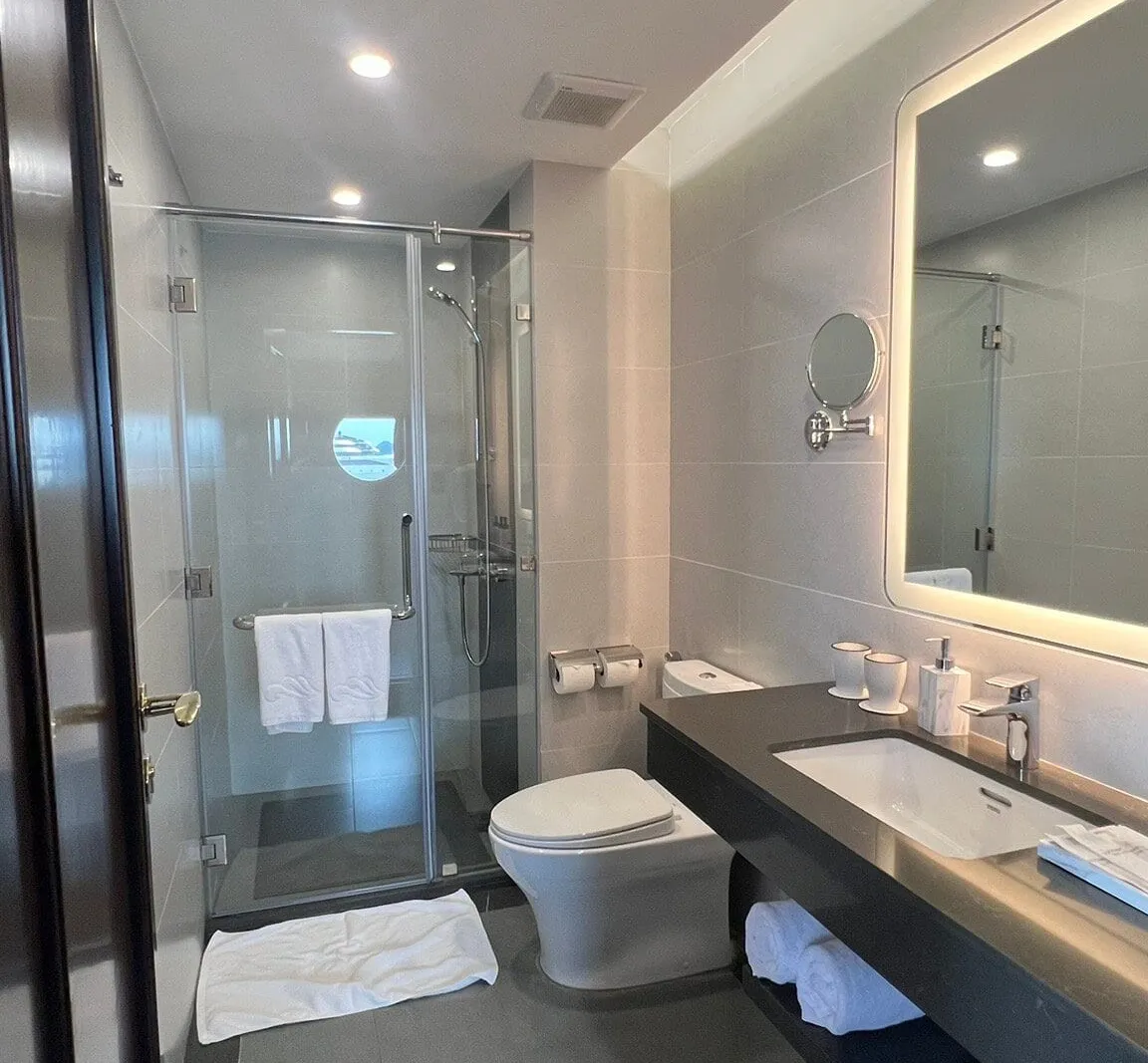 A modern en-suite bathroom with a glass shower enclosure, toilet, backlit mirror and dark vanity with towels, with a round porthole window visible in the shower wall and a wave-like emblem embroidered on the hanging towels.