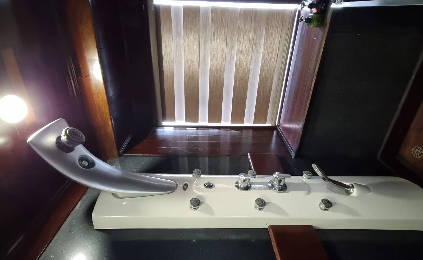 A wide-angle view of a dark wood-paneled bathroom with a white jacuzzi tub fitted with massage jets and a faucet, and a frosted slatted window or door at the back.