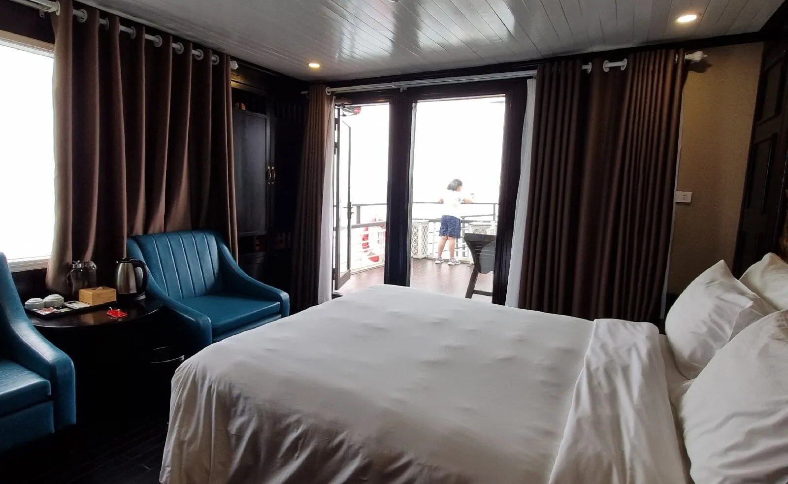 A wide-angle view of a cruise cabin with a white-linen bed, teal armchairs, brown curtains and a gold carved wall panel, with a person standing on the balcony outside the open glass doors in misty, white-grey light.