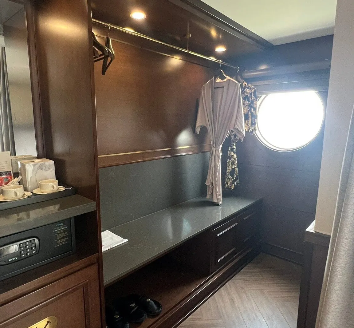 A dark-wood cabin dressing area with a hanging robe, in-room safe, tea cups, slippers on a shelf, herringbone floor and a round porthole window letting in bright light.