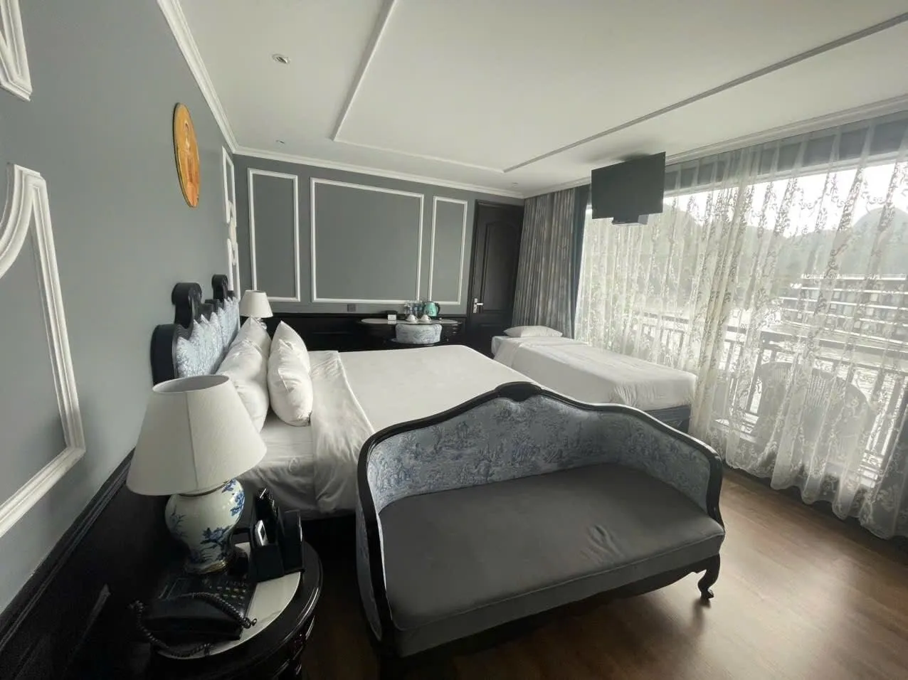 A wide-angle view of a cabin with a double bed, a single bed, a blue-and-white upholstered bench, a porcelain lamp, and a bedside phone, with sheer curtains over a balcony window showing limestone karst hills outside.