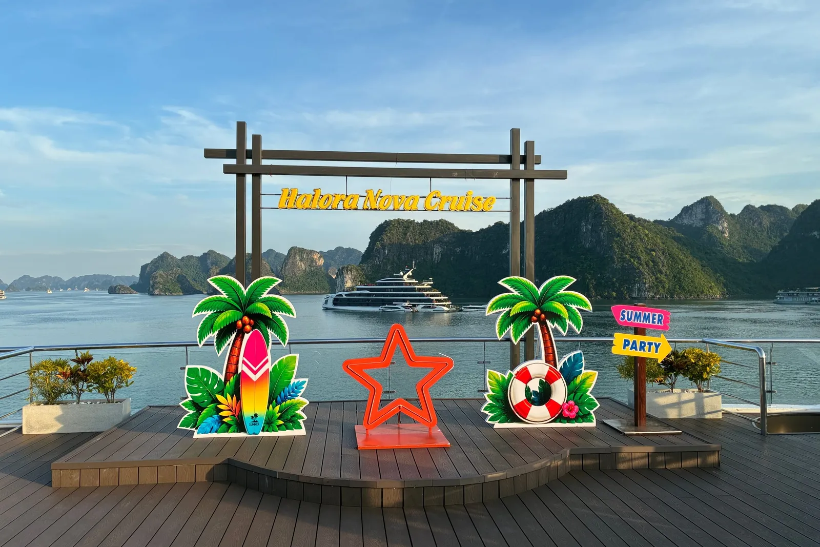 A wooden sun deck with a photo-op set of palm tree, surfboard, star and 'Summer Party' signs under a 'Halora Nova Cruise' sign frame, overlooking limestone karst islands and a distant cruise ship.