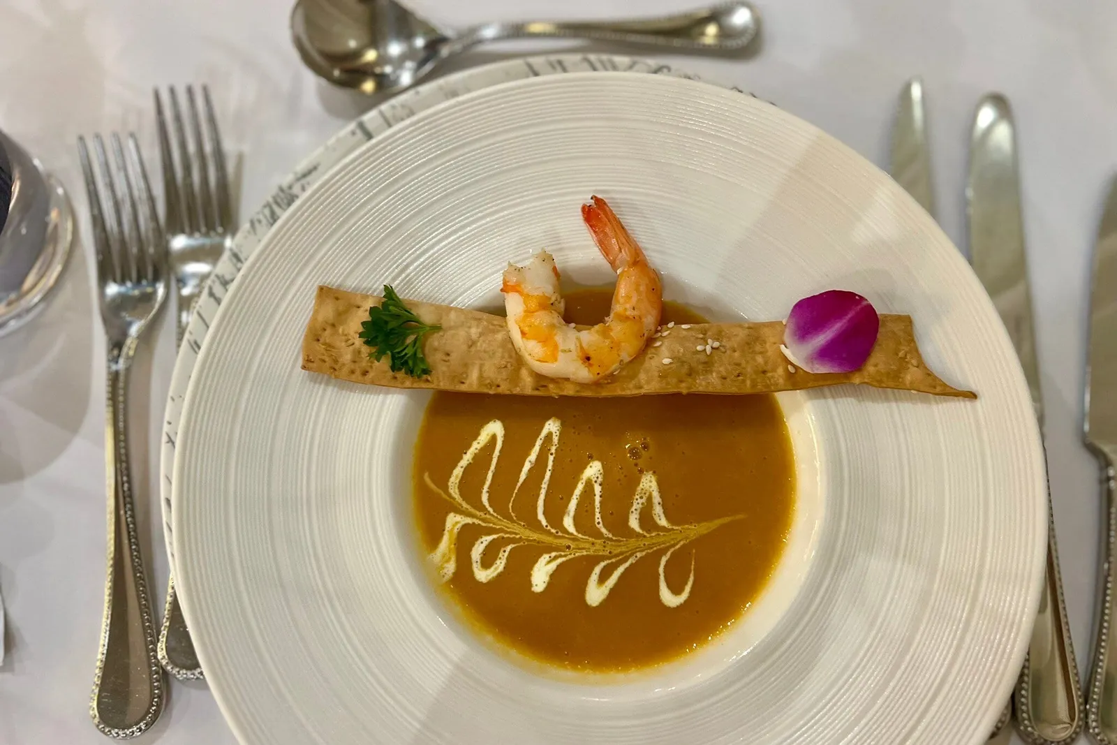 Overhead view of a white bowl of orange bisque-style soup with a cream swirl, topped with a cracker strip holding a shrimp, parsley and an orchid petal, set on a white tablecloth with forks, spoon and knives.