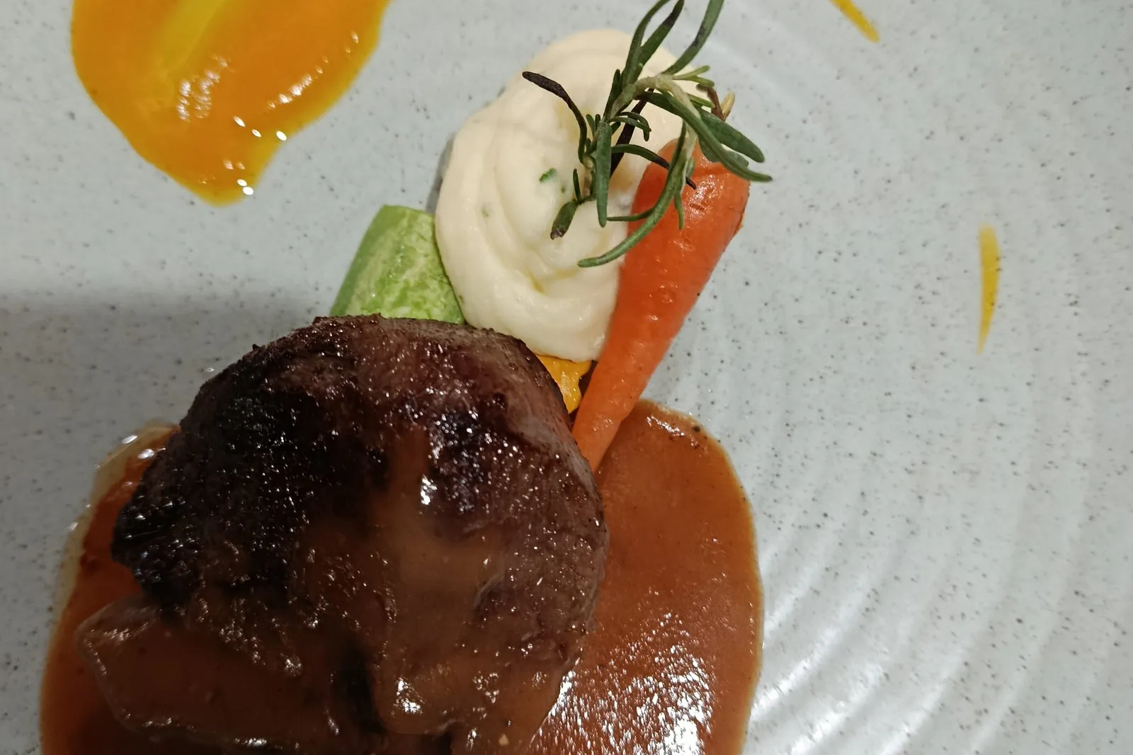 A plated seared beef fillet with brown sauce, mashed potato, a baby carrot, rosemary sprig and orange purée swirl on a speckled grey plate printed with 'Aspira Cruises'.