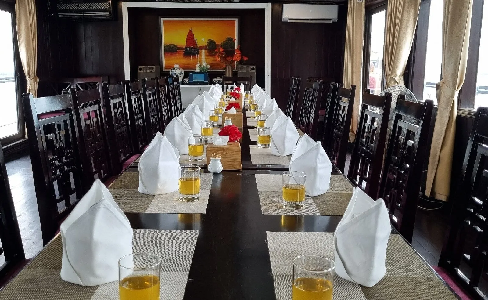 A long dark wooden dining table set with folded white napkins, glasses of orange drink and red flower decorations, flanked by carved dark wood chairs in a cabin with curtained windows and a painting of a junk boat at the far end.