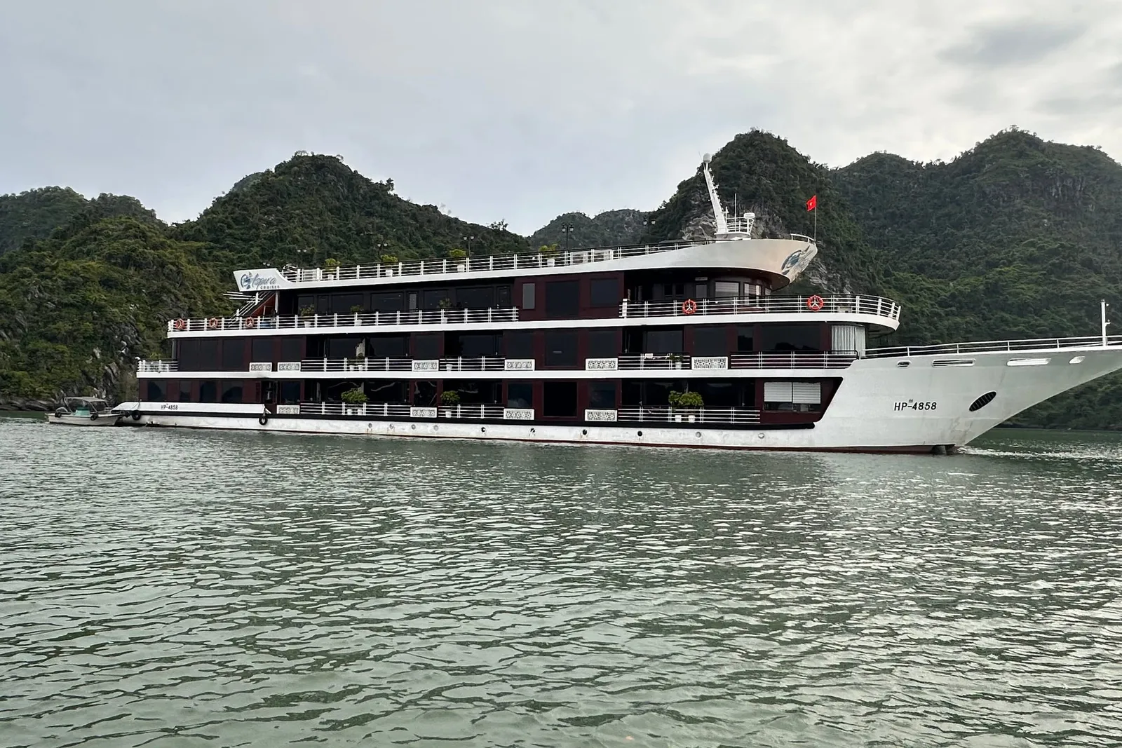 A large white and dark-maroon multi-deck cruise ship with hull number HP-4858 and a red flag sails on green water in front of forested limestone karst hills under an overcast sky.