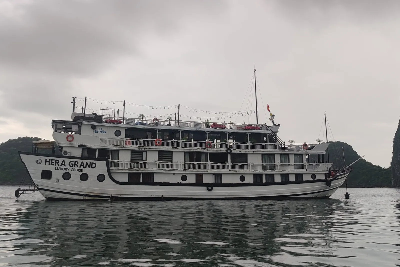 A white multi-deck cruise boat with 'HERA GRAND Luxury Cruise' painted on its hull sits on calm grey water under a heavy overcast sky, with forested karst hills behind it.
