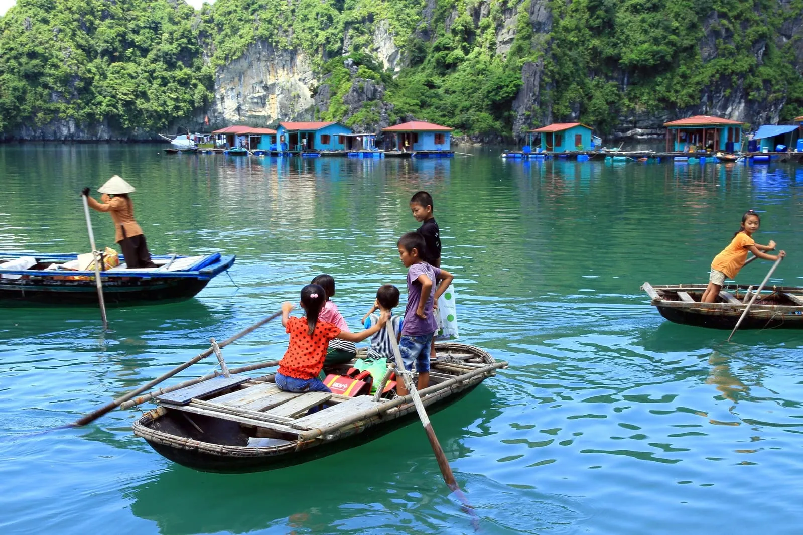 Small rowing boats, one carrying children, between floating houses below a limestone cliff